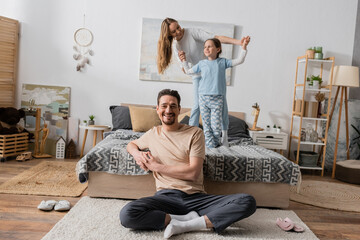Wall Mural - cheerful bearded man sitting on carpet near wife and daughter standing on bed on blurred background.