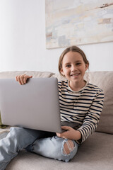 Wall Mural - joyful preteen girl in casual clothes looking at camera and holding laptop while sitting on couch.