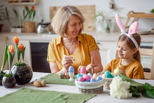 Happy Easter Family Elderly Grandmother And Little Granddaughter With Rabbit Ears Are Preparing For The Holiday To Paint Eggs