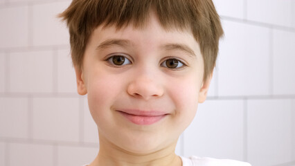 Happy child looks at the camera and smiles. Cute caucasian 6 year old boy in white t-shirt. Close-up portrait. Concept of happiness and good mood. Cheerful kid. Pretty face. Home bathroom background