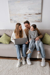 Wall Mural - full length of happy woman sitting with laptop near husband and daughter in living room.