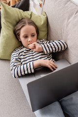 Wall Mural - high angle view of focused preteen girl in casual clothes using laptop while lying on couch.
