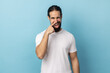 Portrait of crazy handsome man with beard wearing white T-shirt standing and drilling his nose with funny face, winking at camera. Indoor studio shot isolated on blue background.