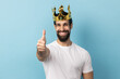 Portrait of smiling joyful man with beard wearing white T-shirt and in gold crown standing looking at camera with toothy smile, showing thumb up. Indoor studio shot isolated on blue background.