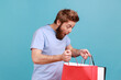 Portrait of attractive bearded man standing, holding red and white shopping bags and looking inside with shocked facial expression. Indoor studio shot isolated on blue background.