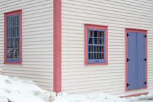 The Exterior Wall Of A White Wooden Cape Cod Clapboard Siding House With A Purple Panel Door And Vintage Glass Window, Black Metal Hinges, Pink Trim Around The Door, And White Snow On The Step. 