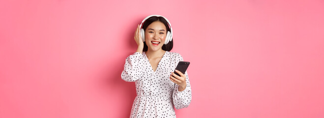 Beautiful asian woman listening music in headphones, using mobile phone, smiling happy at camera, standing over pink background