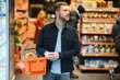 Customer In Supermarket. Man Doing Grocery Shopping Standing With Cart Choosing Food Product Indoors. Guy Buying Groceries In Food Store. Selective Focus, Copy Space.