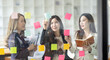 © David - Young creative business Asian woman standing with her colleagues writing new ideas on sticky notes over glass wall, business marketing strategy or User experience ux ui concept.