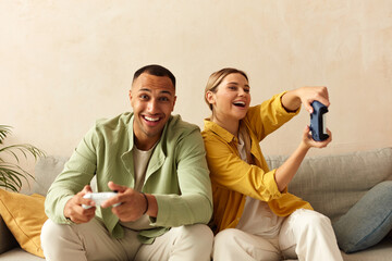 Happy Couple Playing Video Games. Boyfriend And Girlfriend Sitting On Couch In Living Room Enjoying Playing Video Games And Spending Time Together. Enjoying Moment On Weekend Concept