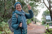 Smiling Senior Woman Walking Outdoors In A Mountain Path Enjoying Freedom And Nature. Cheerful Senior Woman With Backpack Hiking In Park Making Positive Gesture With Fingers