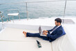 Confident businessman working on laptop computer and talking on mobile phone for global corporate business while travel on luxury private catamaran boat yacht sailing in the ocean on summer vacation.