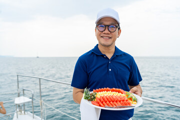 Wall Mural - Portrait of Happy Asian man waiter holding fresh fruit on serving dish for serving to passenger tourist travel on luxury catamaran boat yacht on summer vacation. Cruise ship service occupation concept