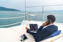 Caucasian Businessman Working On Laptop Computer And Talking On Mobile Phone For Global Corporate Business While Travel On Luxury Private Catamaran Boat Yacht Sailing In The Ocean On Summer Vacation.