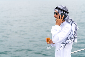 Wall Mural - Arab businessman talking on mobile phone with global corporate business while travel on private catamaran boat yacht sailing in the ocean. Arabic man enjoy luxury outdoor lifestyle on summer vacation.