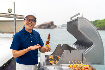 Asian man chef cooking grilled barbecue and seafood on stove for serving to passenger tourist travel on luxury private catamaran boat yacht on summer vacation. Cruise ship service occupation concept.