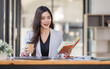 © David - Asian Businesswoman having a tea, sitting on desk in workplace, writing down notes, opened laptop in document the financial report, business plan investment, finance analysis concept.