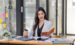 © David - Asian Businesswoman having a tea, sitting on desk in workplace, writing down notes, opened laptop in document the financial report, business plan investment, finance analysis concept.