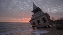 Mysterious Vintage Flooded Leaning Wooden House Or Chapel Washed By Sea Waves, At Coastline On Pink Sunset. Enigmatic Rickety Building Sinking In Water On Sandy Beach Near Breakwater On Sunrise.