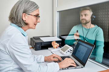 Wall Mural - Experienced female audiologist doing hearing test to male patient in soundproof audiometric booth with audiometer in hearing clinic. Audiometric testing