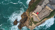 Aerial View Of An Old Lighthouse On A Cliff With A Fortress On The Coast Of The Atlantic Ocean In Nazare Town, Portugal. High Quality Photo