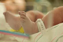Close Up On The Tiny Feet Of A Newborn Baby Girl, Laying In A Hospital Bed As Her Mother Rests In The Background.