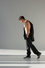 Full Length Of Shirtless Man With Hand In Pocket Of Black Trousers Walking In Light On Grey Background.