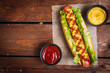 Hot dog with mustard, ketchup and green salad in bun, served with potato chips on a rustic wooden board. Fast food and delicious.