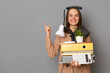 © sementsova321 - Portrait of funny young pretty woman wearing beige jacket holding documents and her office stuff posing with phone on her head isolated over gray background, showing advertisement area.