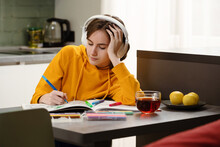 Teenager Girl Sits At The Table, Paints Images And Listens Music While Drinking Tea On Kitchen. Headphones On Her Head. Recreation And Relaxation Hobby