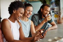 Group Of Smiling Friends Looking At Cellphone Photos Over Coffee