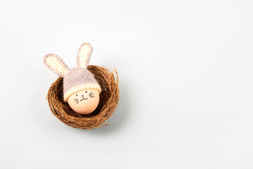 Easter egg with funny face and gray crocheted hat with bunny ears in a decorative nest of sisal on a gray background. Happy Easter concept. Greeting card.