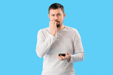 Handsome Man With Mobile Phone Biting Nails On Blue Background