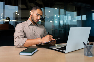 Serious concentrated businessman writing document while sitting in office using laptop for online learning, hispanic man in glasses listening to video call report at workplace.