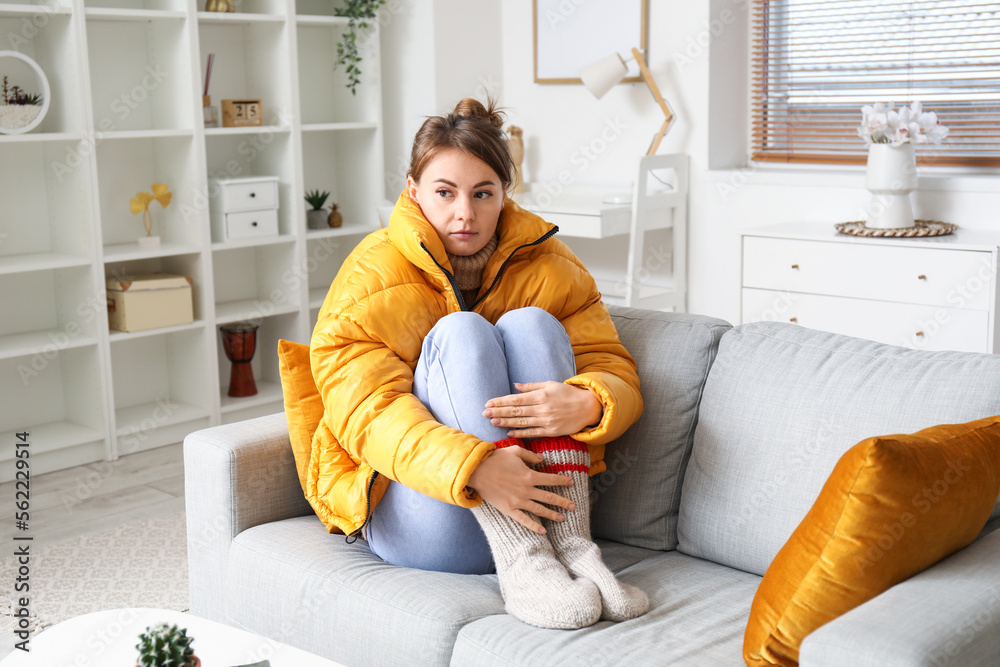 Frozen young woman in down jacket sitting on sofa at home