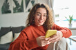 © insta_photos - Young smiling woman wearing glasses holding smartphone using cellphone modern technology, looking at mobile, checking cell phone apps, texting, browsing internet for shopping sitting at home.