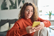 Young Smiling Woman Wearing Glasses Holding Smartphone Using Cellphone Modern Technology, Looking At Mobile, Checking Cell Phone Apps, Texting, Browsing Internet For Shopping Sitting At Home.