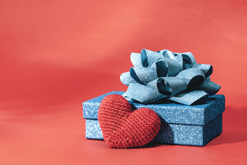 Wall Mural - Knitted heart Blue sparkling gift box with ribbon bow on red background. Gift or holiday concept. Mothers Day, Fathers birthday wedding or St Valentines day with copy space. Minimal banner