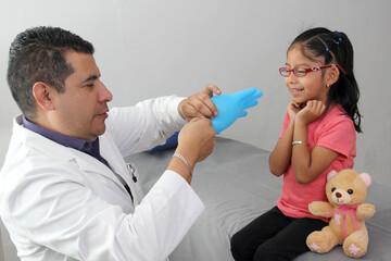 Wall Mural - Pediatrician doctor and patient 4-year-old Latino brown-haired girl play in the office with a glove as a balloon with a happy face so that she relaxes and is not nervous in the medical consultation