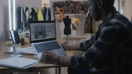 African American fashion designer develops clothes design in 3D modeling program on laptop using digital tablet and stylus. He works in atelier workshop. Fashion and technologies in business concept.