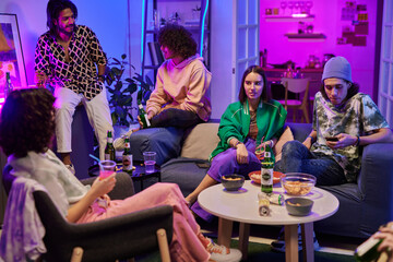 Group of youthful friends in casualwear having drinks while relaxing in living room lit by neon light and enjoying home party