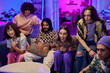 © pressmaster - Large group of happy young friends sitting on couch in living room lit by neon light and watching broadcast at home party on weekend