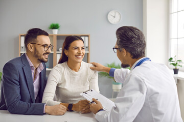 Joyful young family couple planning pregnancy and visiting doctor. Medical specialist sitting at desk with happy man and woman, talking about IVF treatment, and reassuring them