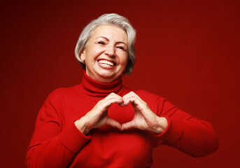 senior grey-haired woman wearing red sweater smiling in love doing heart symbol shape with hands.