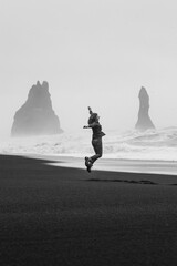 Wall Mural - Happy tourist jumping on black northern monochrome beach scenic photography. Picture of person with cliffs on background. High quality wallpaper. Photo concept for ads, travel blog, magazine, article