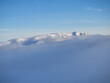 Aerial view of distant mountain peaks above clouds in clear sunny blue sky.