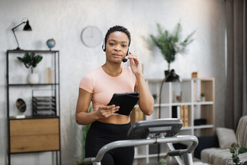 Portrait of active and dynamic young pretty businesswoman with headset and tablet doing sport fitness at home running on treadmill indoor at night. Tone your body. Perfect shape.