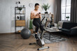 © Sofiia Shunkina - Concentrated african sports women in sportswear tapping on her mobile phone or watching online class, cycling bike at home. Cardio training, exercising legs, cardio workout indoors.