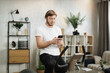 © Sofiia Shunkina - Portrait of focused young male wearing sportswear using exercise bike typing message on phone. Home fitness workout sporty man training on smart stationary bike indoors.
