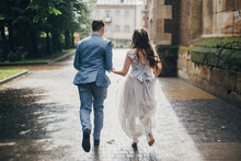 Stylish Bride And Groom Running On Background Of Old Church In Rainy Street. Provence Wedding. Beautiful Emotional Wedding Couple Having Fun In Rain In European City.